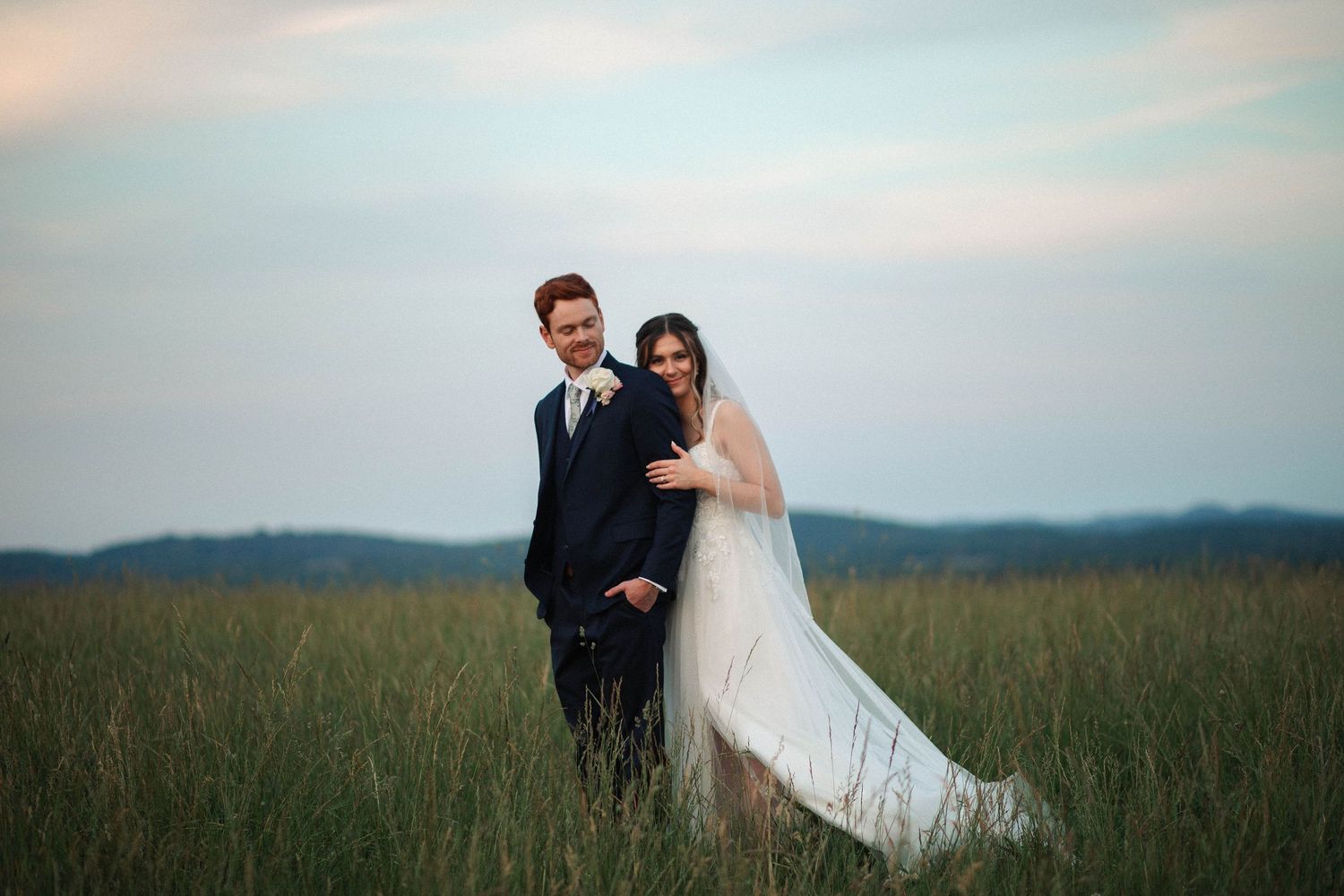 A bride and groom pose together in a lush green field at dusk, bride in white gown with veil, groom in navy suit.