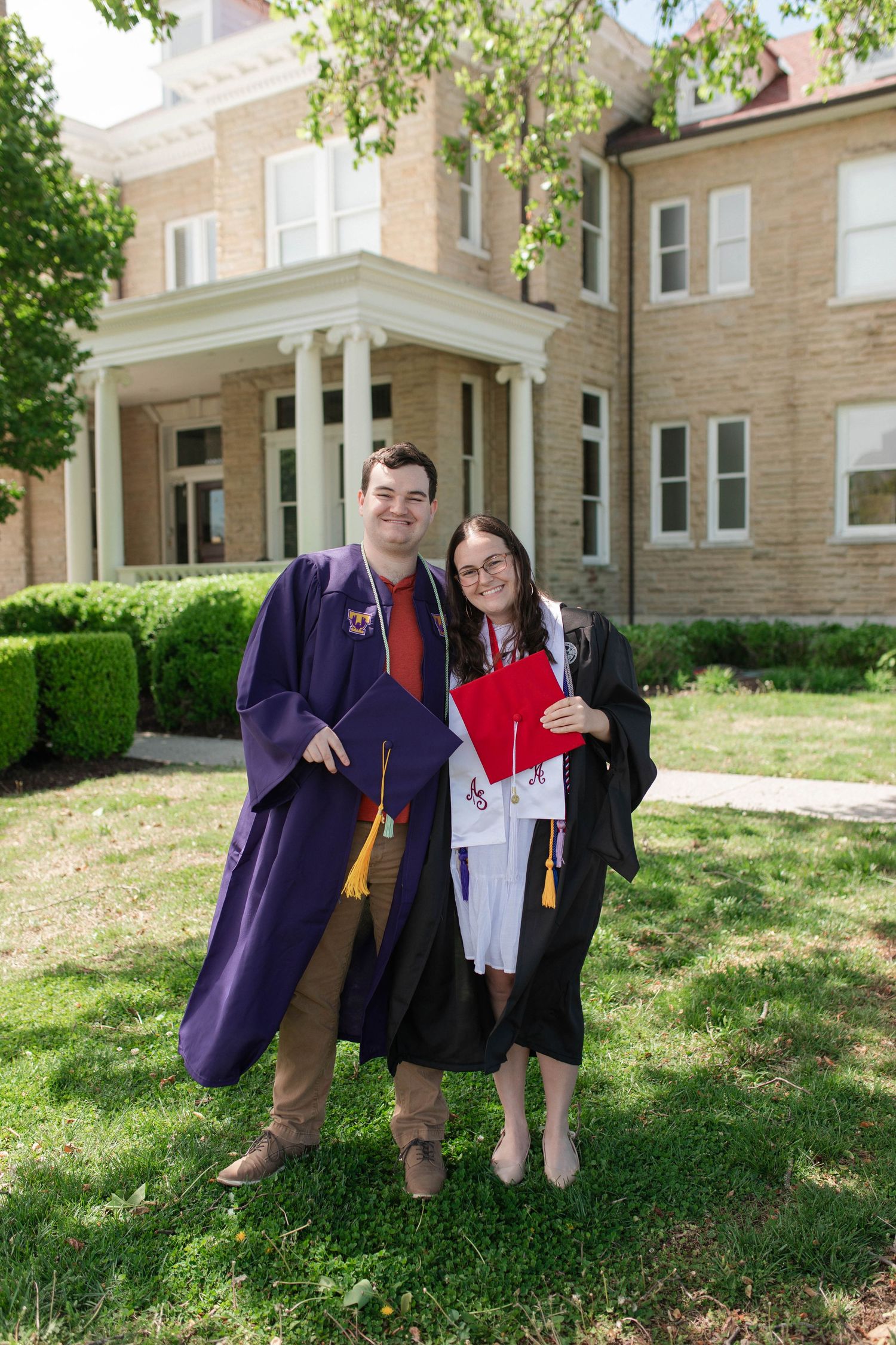 Two graduates in purple and black gowns smiling together outdoors in front of a brick building on graduation day.