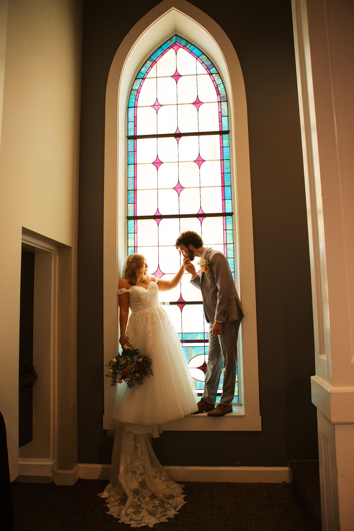 Bride and groom share a romantic moment by a stunning stained glass church window on their wedding day.