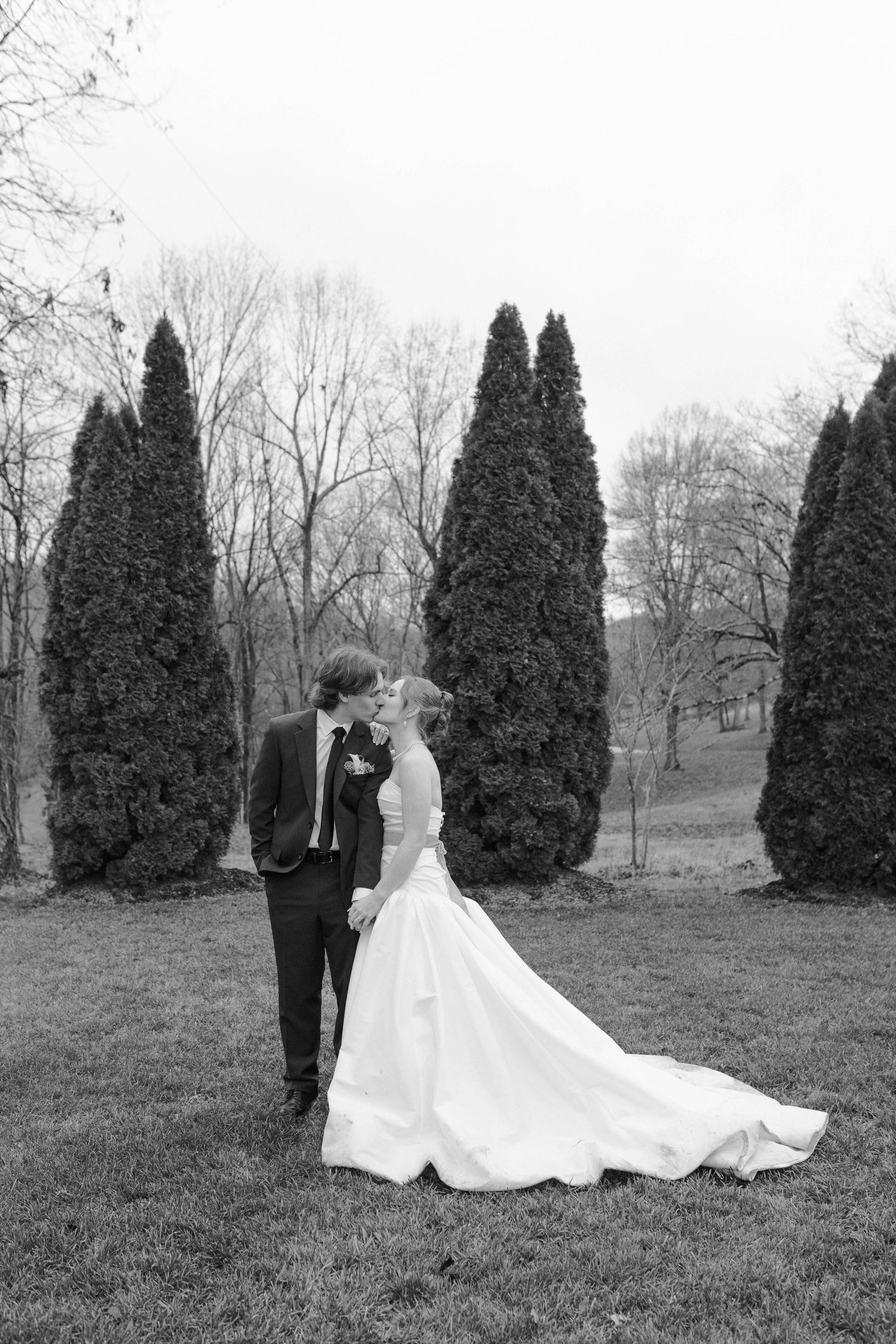 Black and white photo of bride and groom kissing near tall evergreen trees after eloping in Nashville.