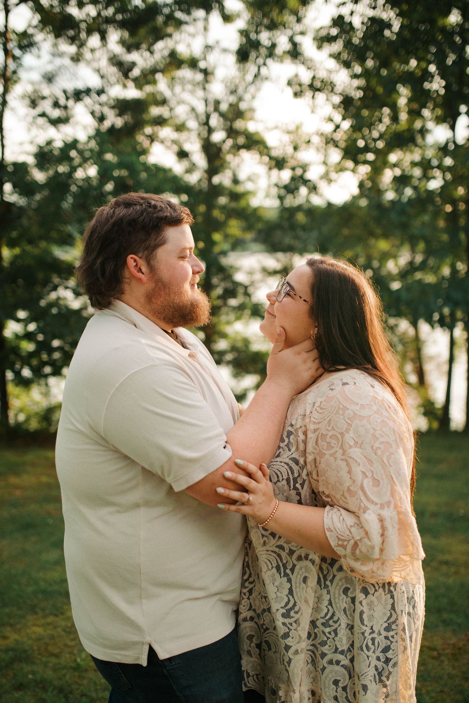 A couple shares a tender moment outdoors, man gently cupping womans face, surrounded by lush green trees at golden hour.