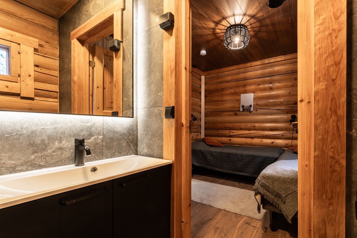 Rustic log cabin bathroom with modern white sink vanity and mirror, opening to a cozy wood-paneled bedroom.