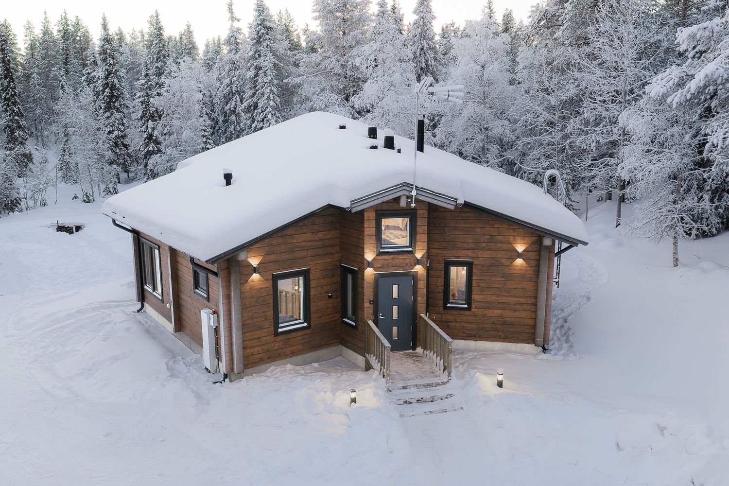 Snow-covered wooden cabin with warm exterior lighting surrounded by frost-covered pine trees in winter.