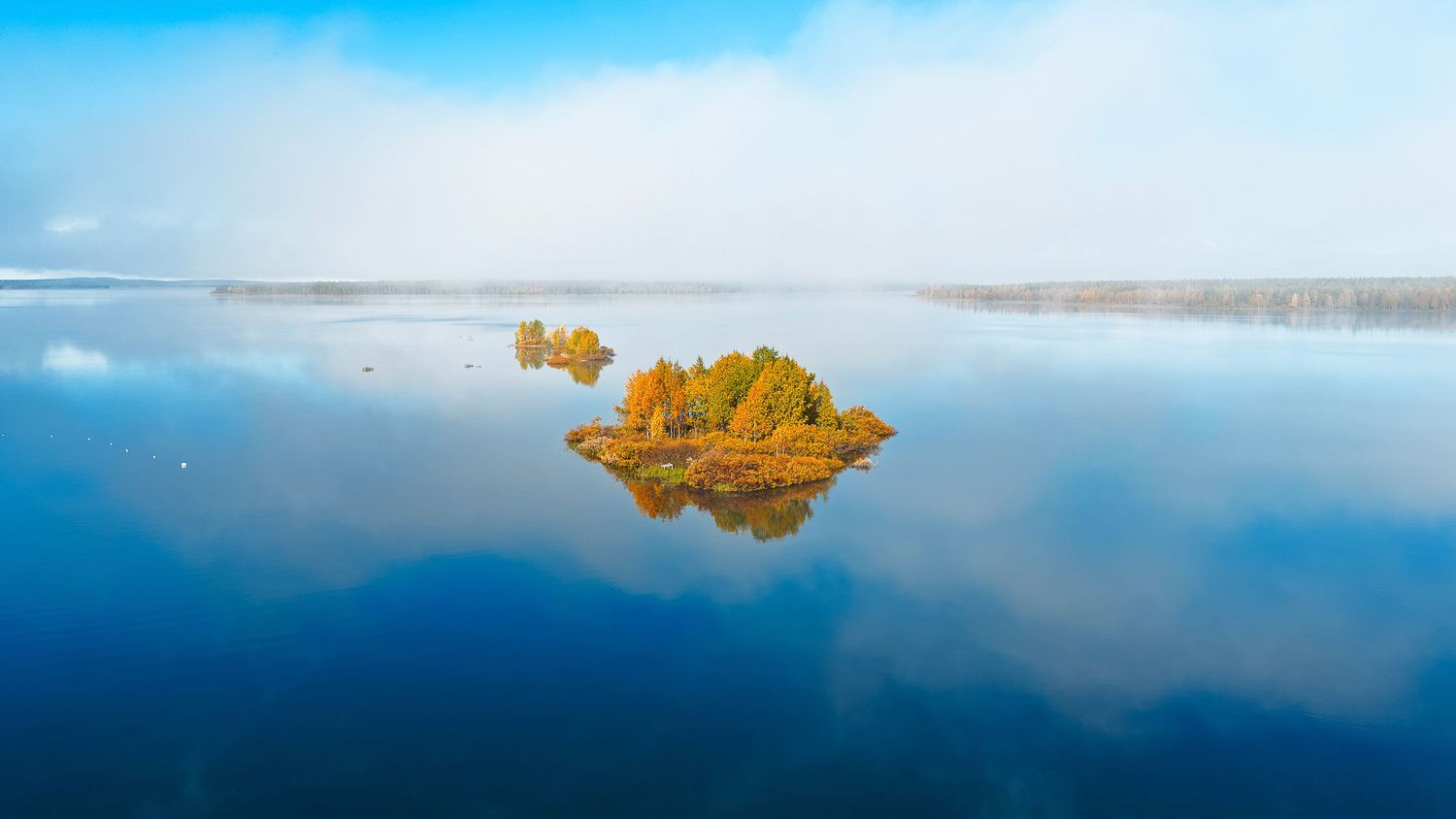 Aerial view of a small island with autumn trees reflecting on a calm misty lake under a blue sky.