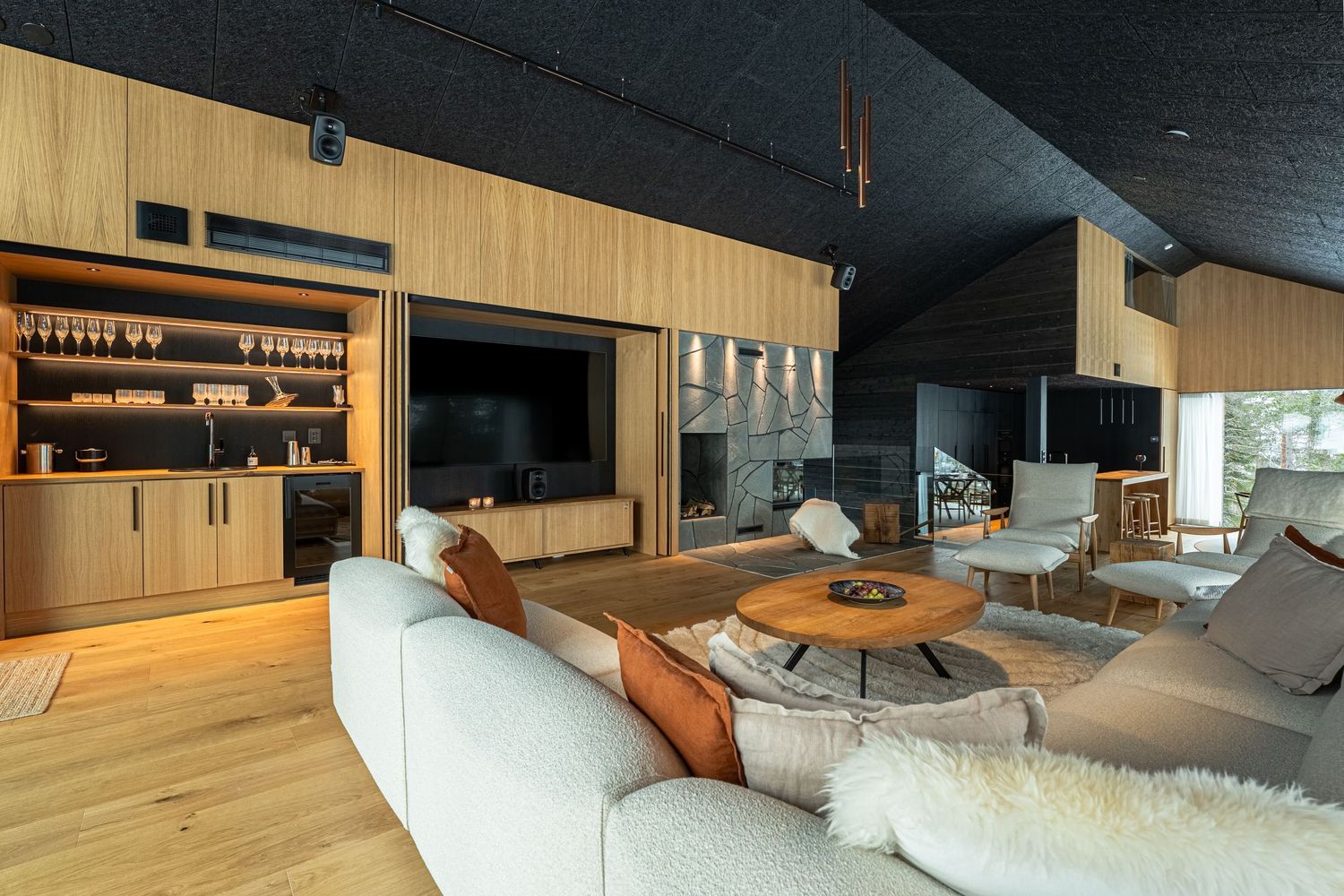 Luxurious modern living room with light wood paneling, built-in bar, large TV, and plush white sofas around a round coffee table.