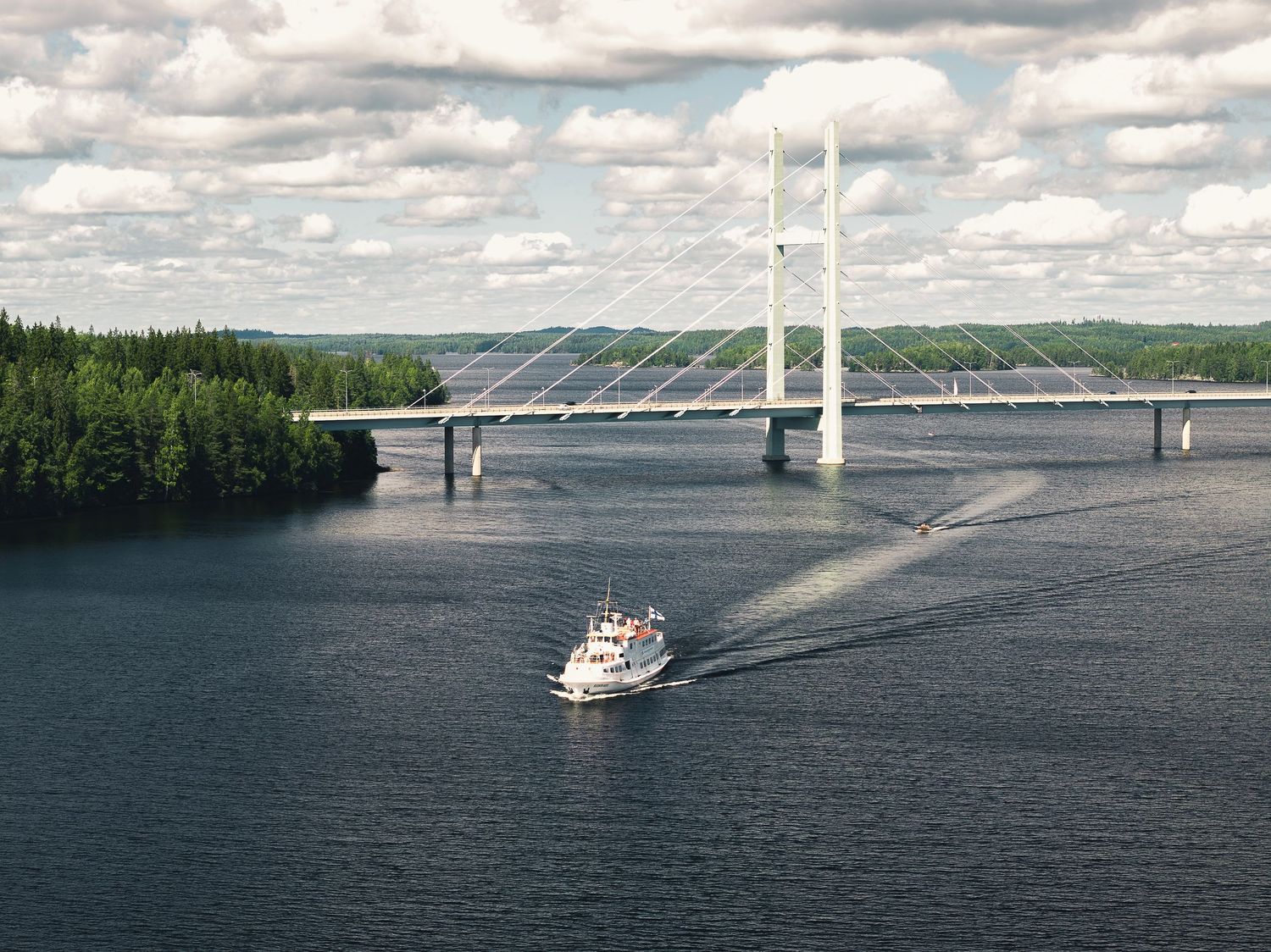 A white ferry boat cruises past a modern cable-stayed bridge over a calm blue lake surrounded by lush forests.