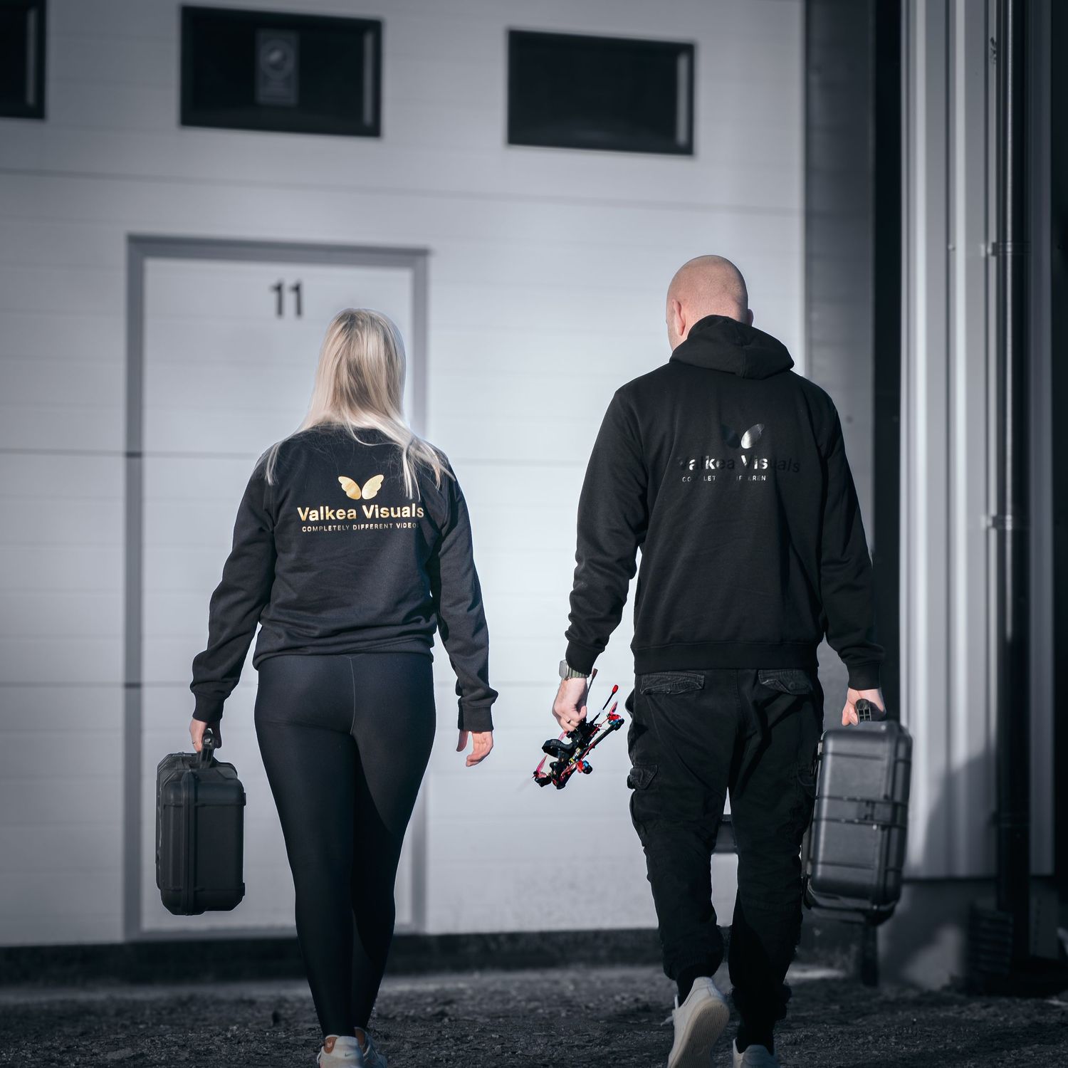 Two people carrying camera equipment cases walking toward building door, one wearing Valkas Visuals hoodie.