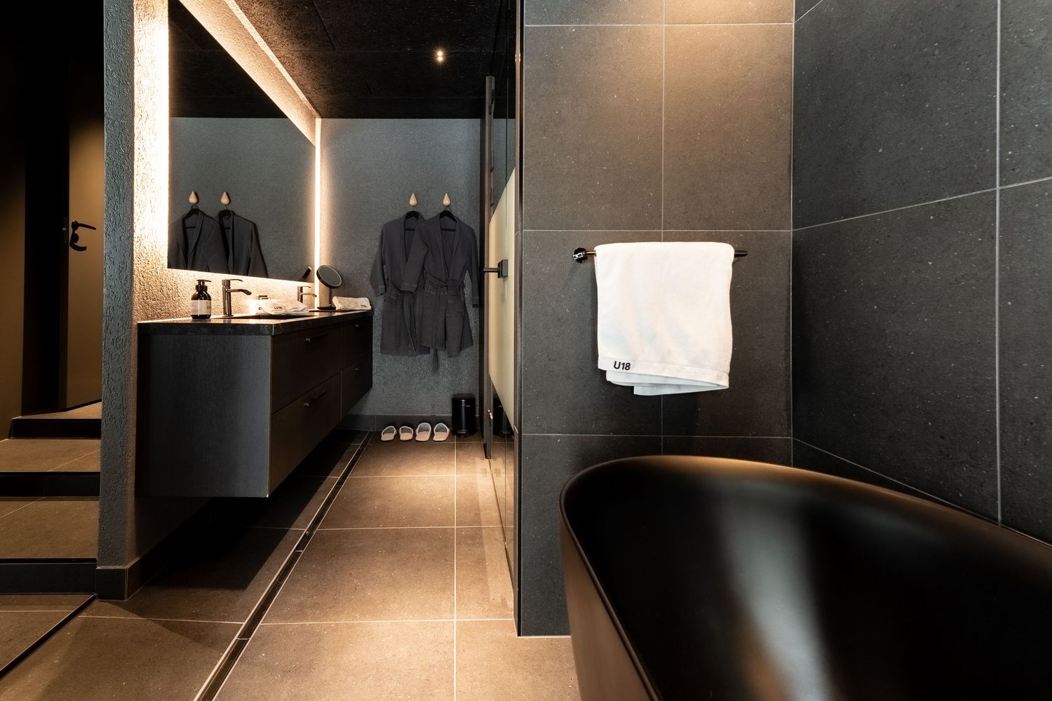 Elegant dark bathroom featuring a black freestanding bathtub, dual vanity with lit mirror, and hanging robes.