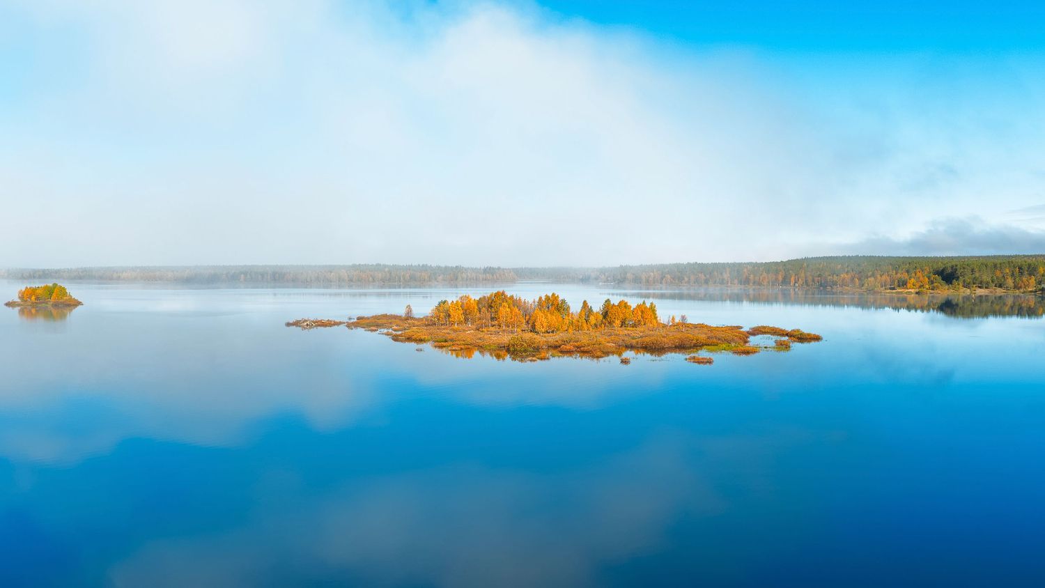 Aerial view of a small island with autumn trees on a calm reflective lake under a misty blue sky.