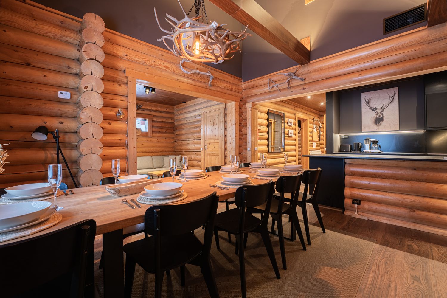 Rustic log cabin dining room with long wooden table, black chairs, antler chandelier, and stag artwork on the wall.