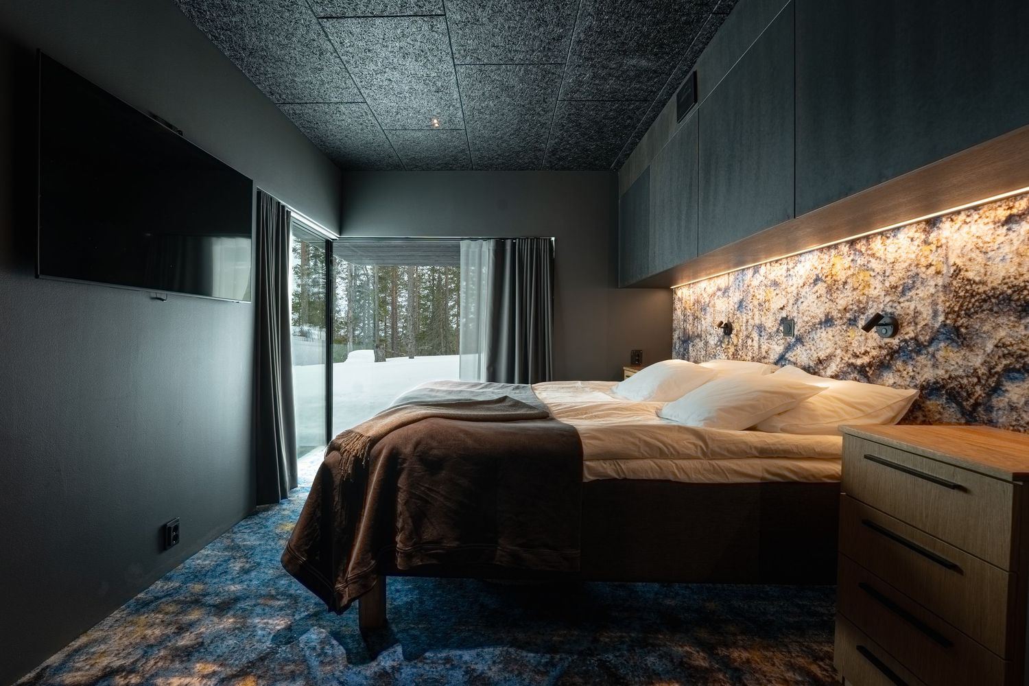 Cozy dark bedroom with textured headboard wall, ambient lighting, blue patterned carpet, and snowy forest view.