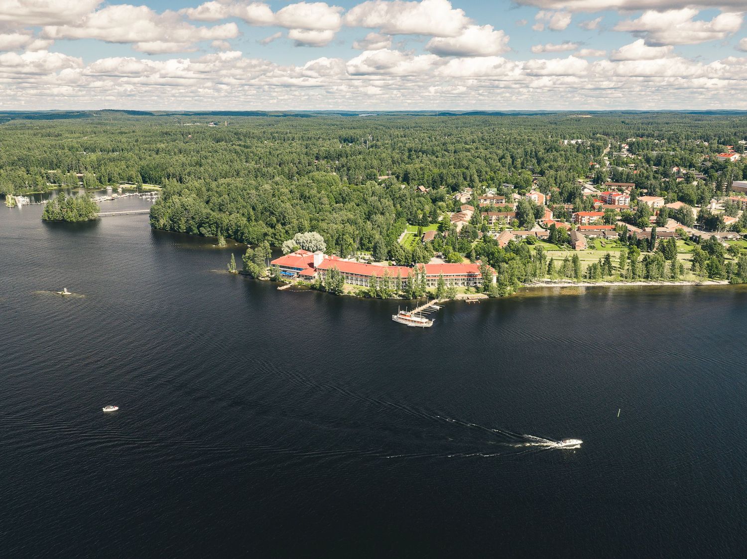 Aerial view of a lakeside resort surrounded by lush green forest with boats on calm blue water in summer.