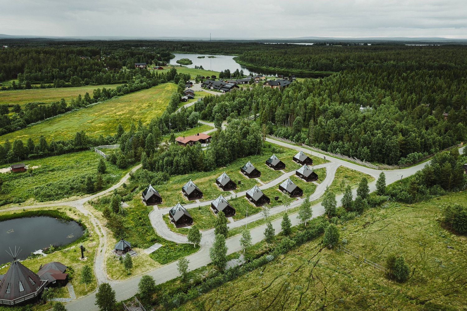 Aerial view of a Scandinavian holiday village with wooden cabins surrounded by lush green forest and a river in the background.
