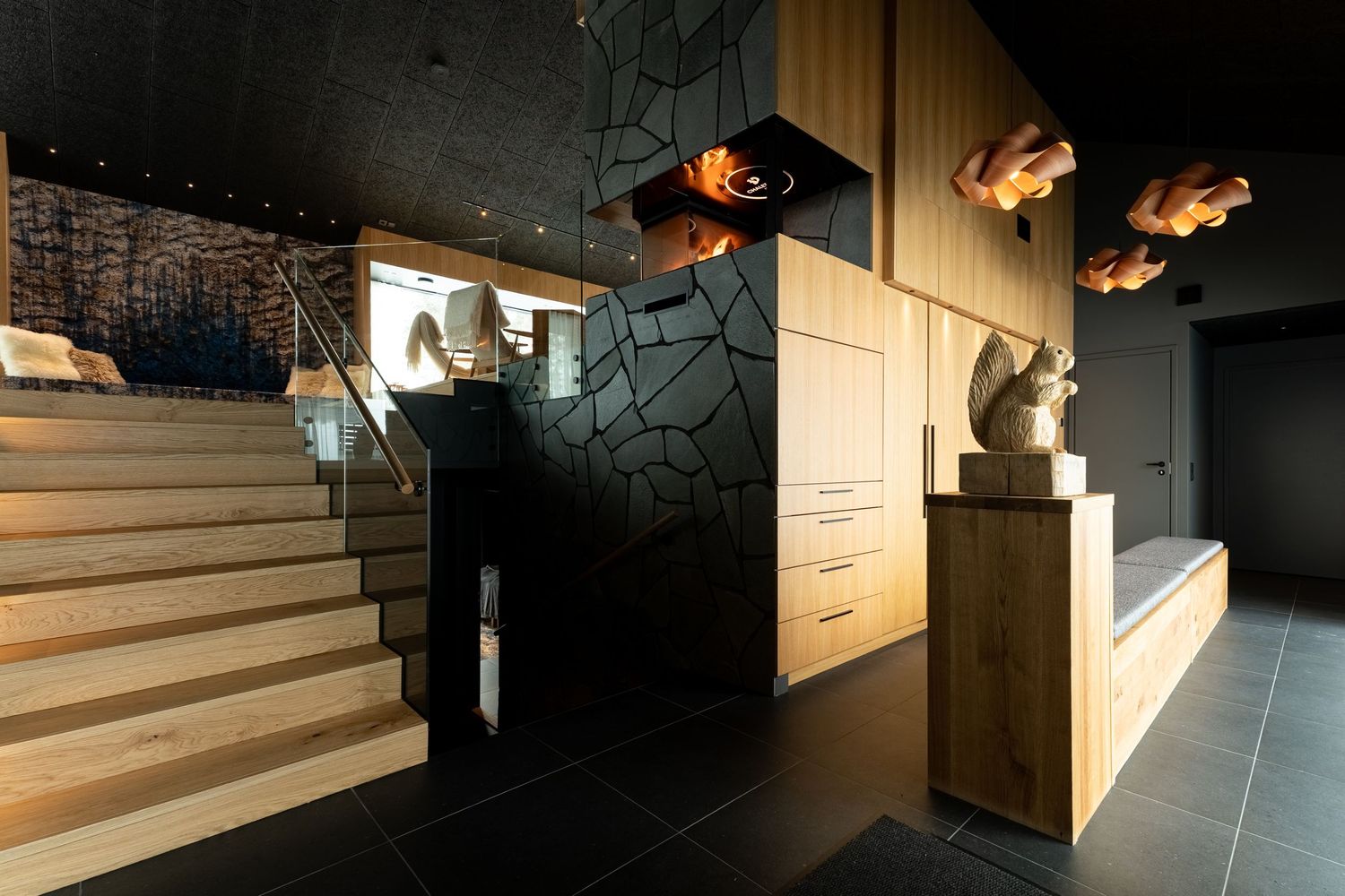 Modern hotel lobby with wooden staircase, geometric black wall panels, sculptural art pieces, and dark ceiling.