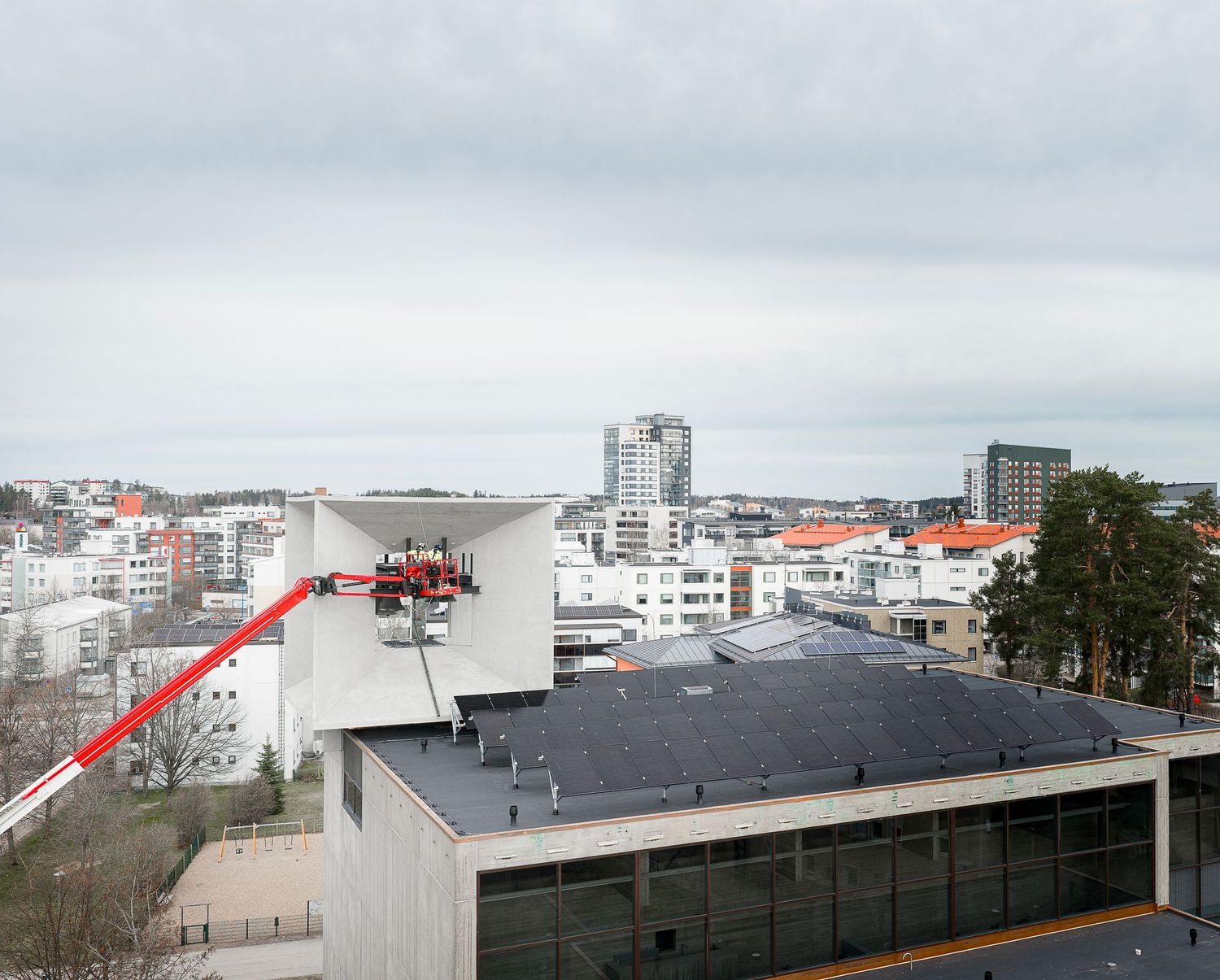Aerial view of a city with a red crane lift on a rooftop, surrounded by urban buildings under an overcast sky.