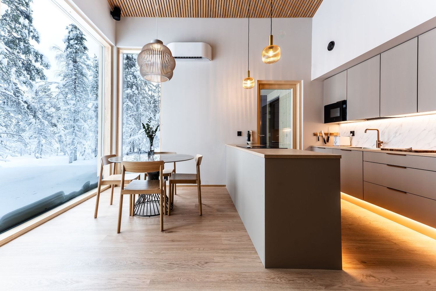 Bright Nordic kitchen featuring taupe cabinetry, warm pendant lighting, and panoramic snowy forest window.