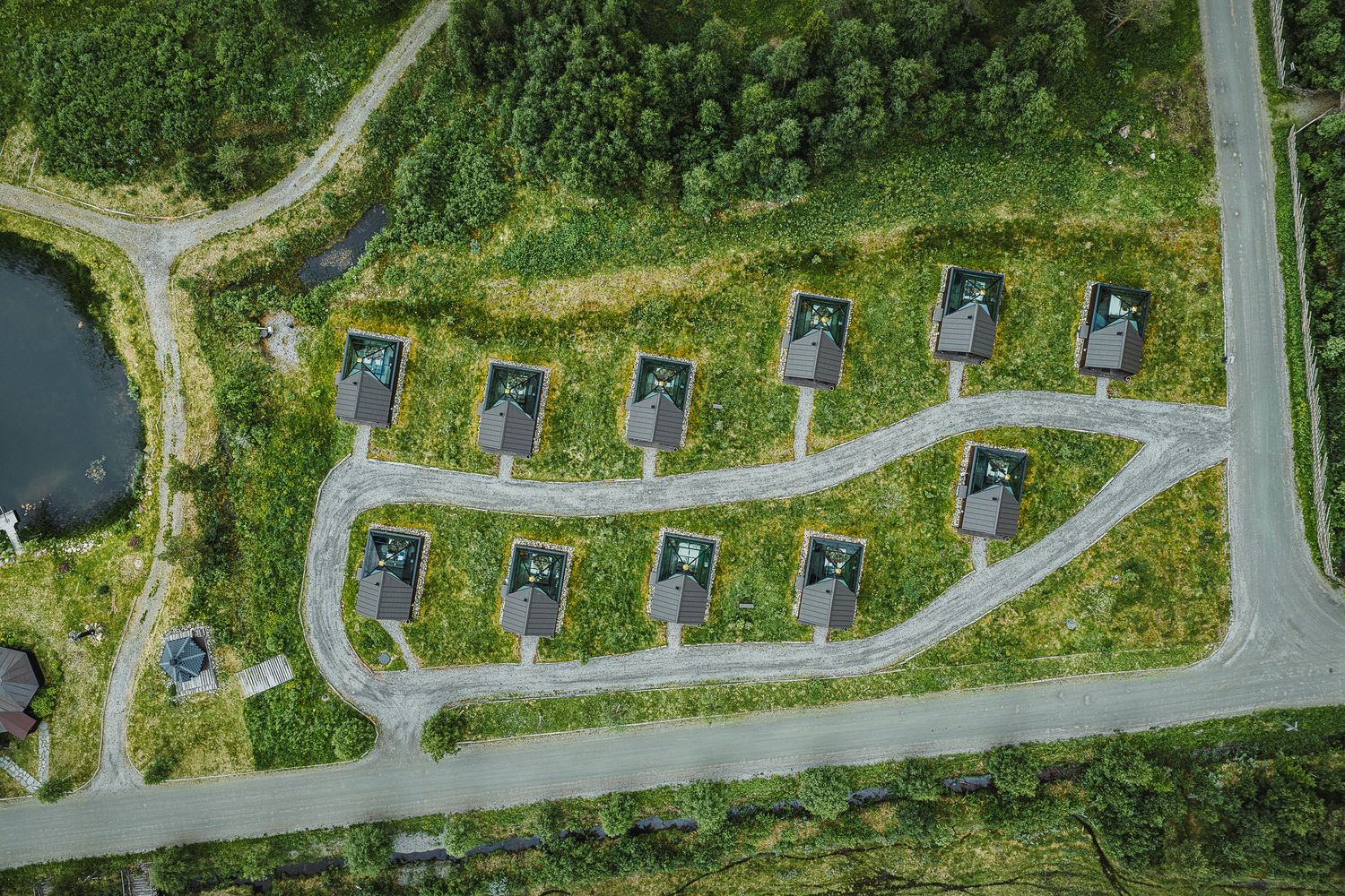 Aerial view of a small cabin resort with rows of dark-roofed cottages along a winding path beside a green pond.