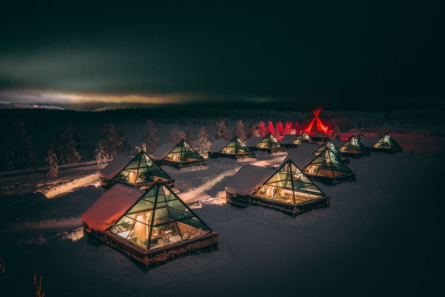 Aerial view of illuminated glass pyramid glamping cabins in snowy Lapland landscape at night.