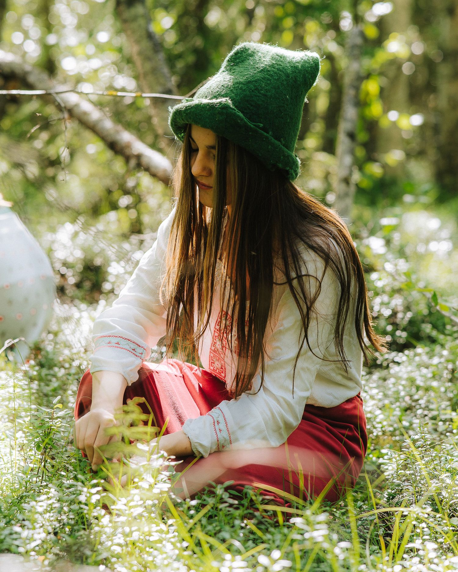 A young girl in a green hat and red skirt sits among white wildflowers in a sunlit forest.