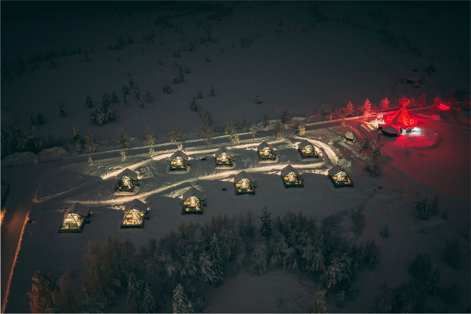 Aerial night view of snow-covered glamping resort with illuminated glass igloo cabins arranged in a winter landscape.