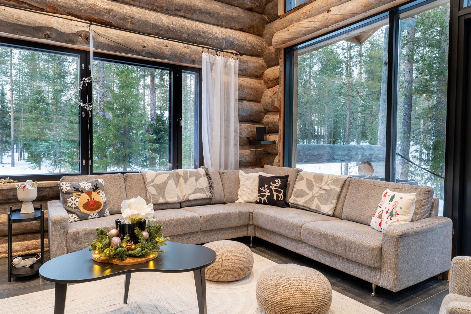 Cozy log cabin living room with gray sectional sofa, decorative pillows, and snowy forest views through large windows.