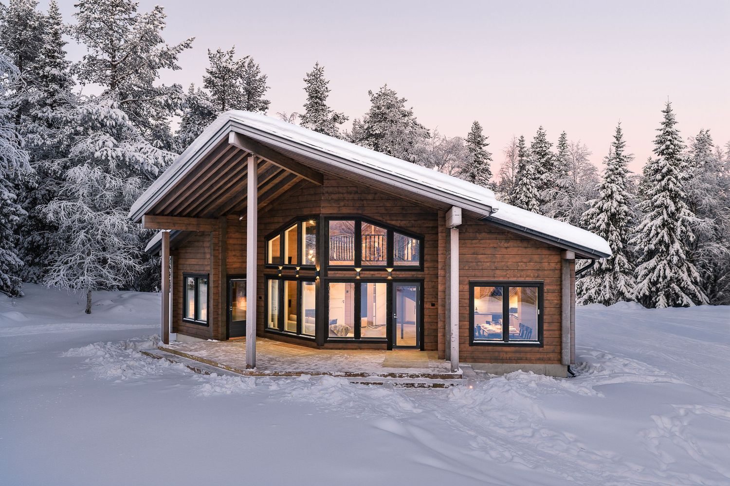 Modern log cabin with large windows glowing warmly at dusk surrounded by snow-covered trees in winter.