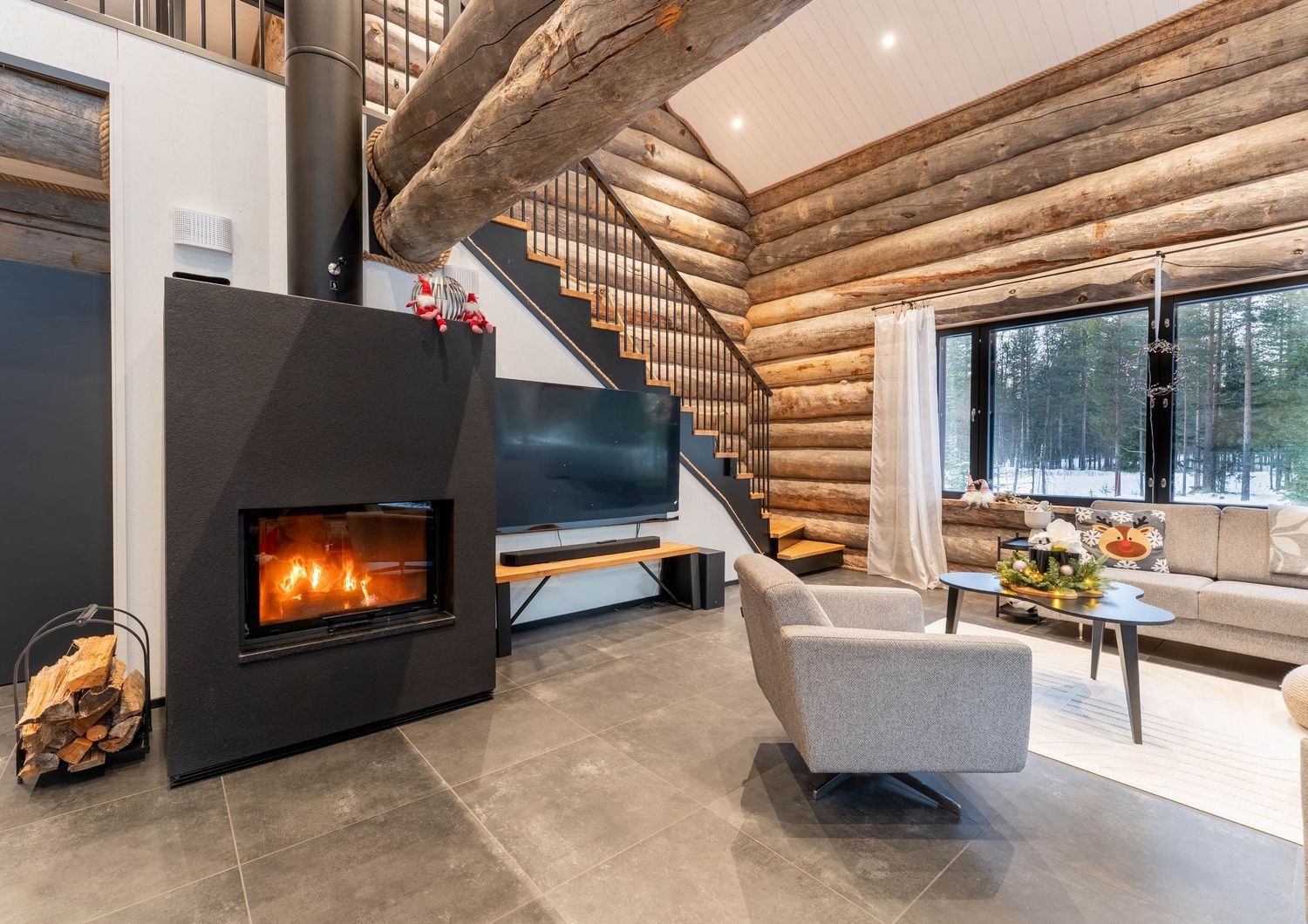Rustic log cabin interior featuring modern fireplace, spiral staircase, TV unit, and snowy woodland window views.