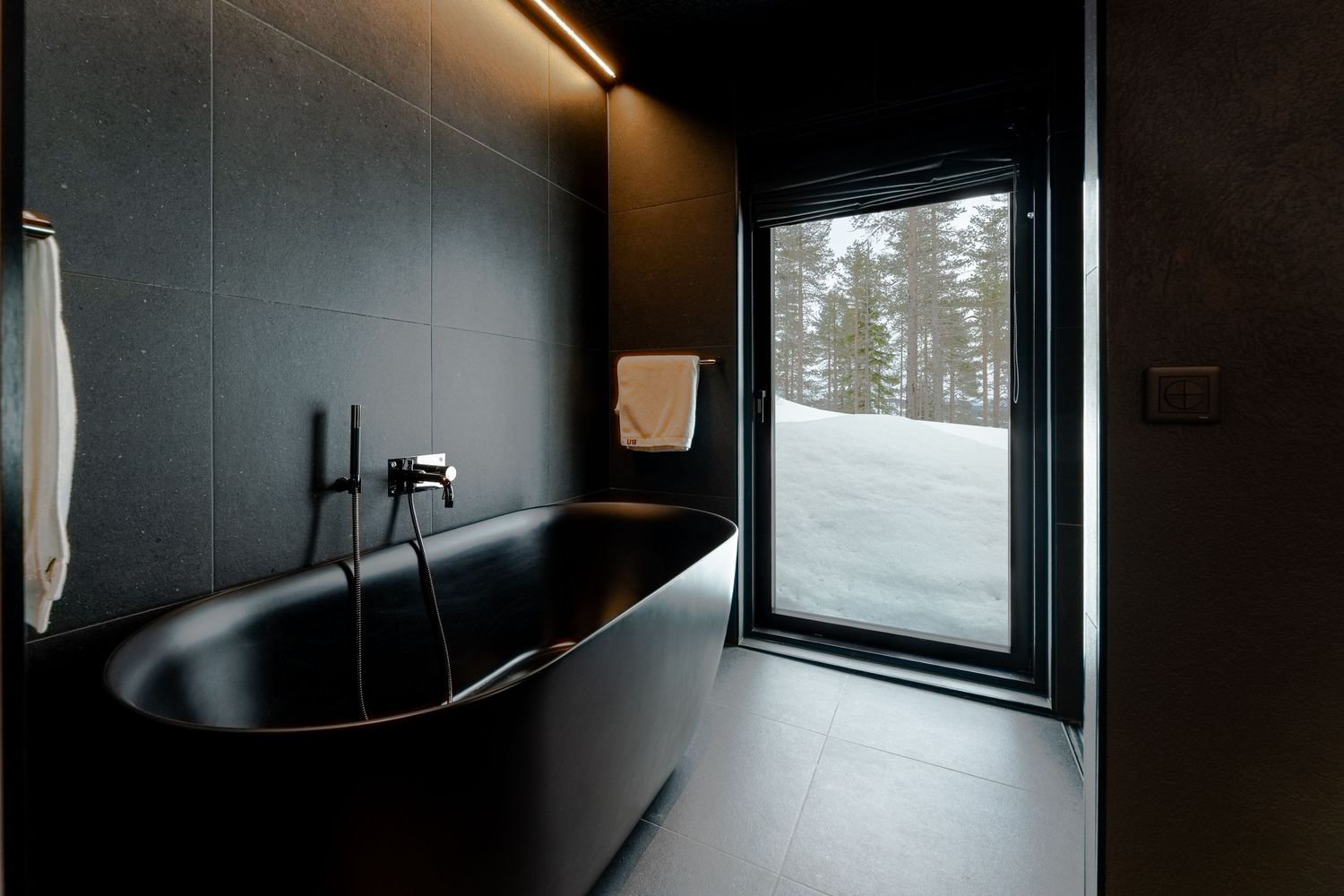 Minimalist black bathroom with freestanding soaking tub and large window overlooking a snowy winter forest landscape.