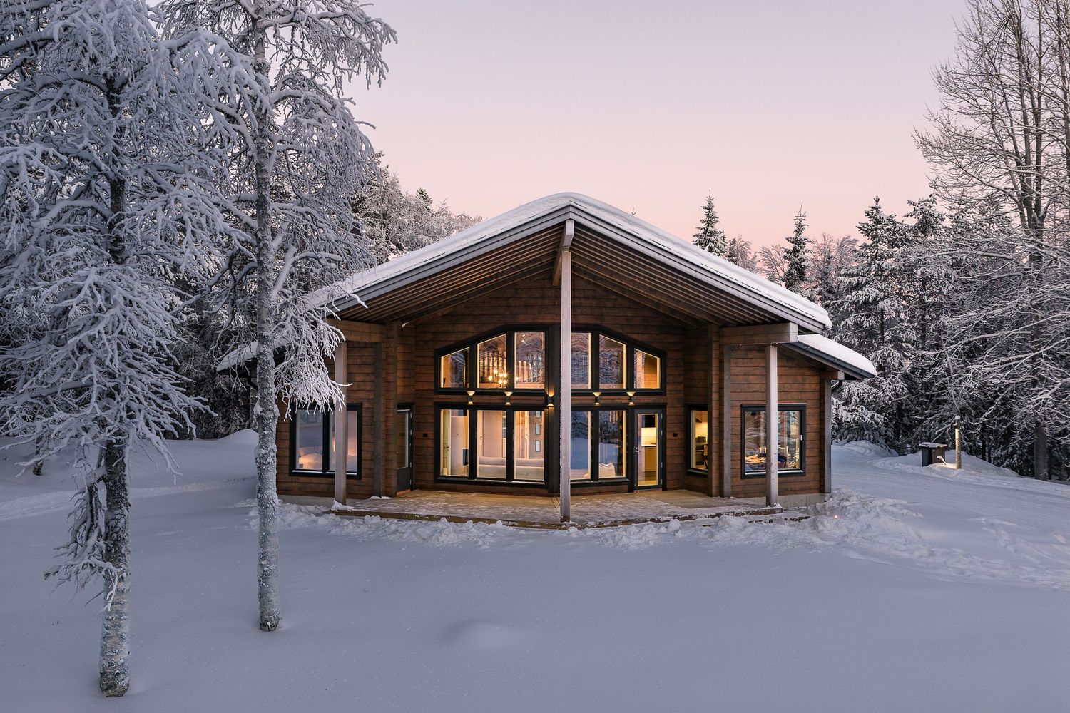 Cozy illuminated log cabin with large panoramic windows surrounded by snow-laden trees at winter twilight.
