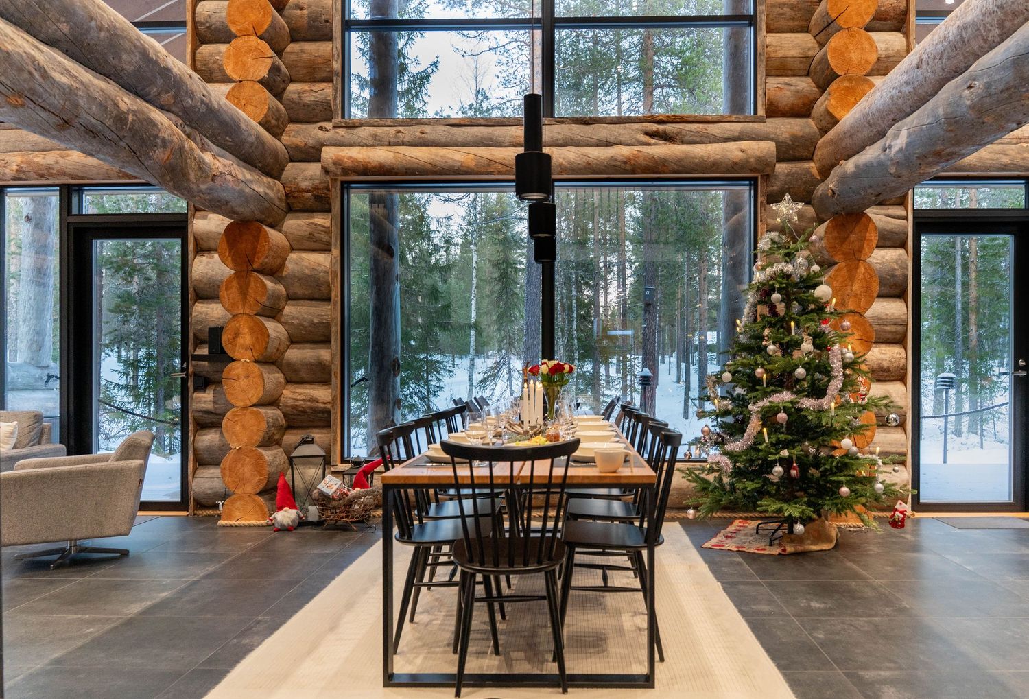 Elegant log cabin dining room with long wooden table, black chairs, Christmas tree, and floor-to-ceiling winter views.