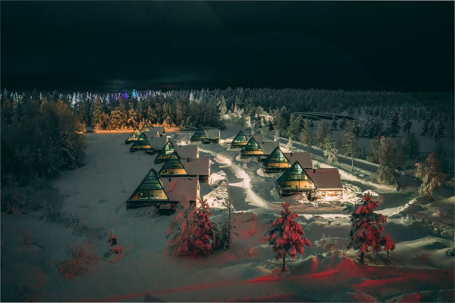Aerial night view of illuminated A-frame chalets surrounded by snow-covered trees in a winter resort village.