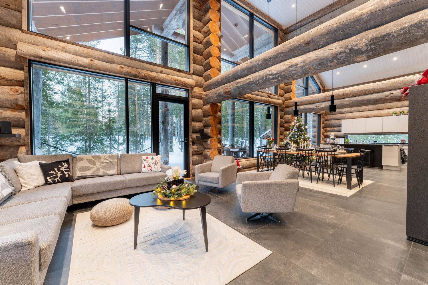 Spacious log cabin great room with high ceilings, sectional sofa, Christmas tree, and panoramic snowy forest windows.