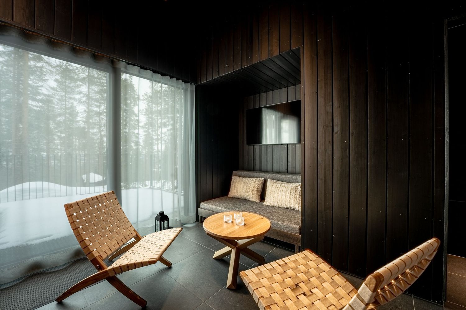 Dark wood cabin lounge with woven leather chairs, small table, sofa nook, and snowy forest views through sheer curtains.