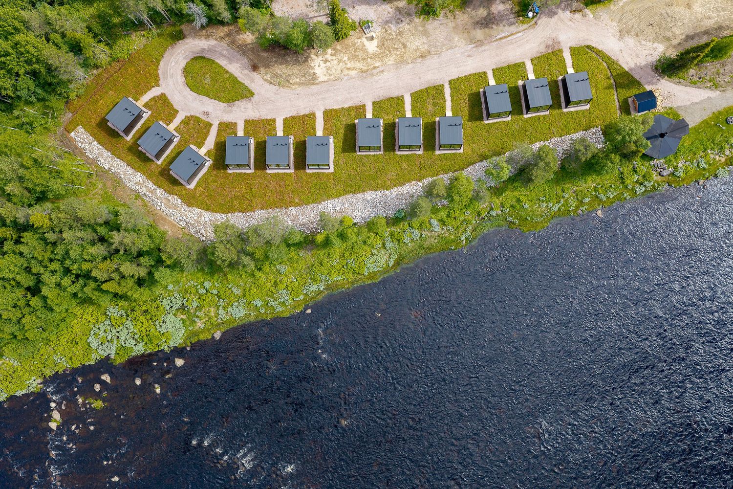 Aerial view of solar-paneled cabins along a scenic riverbank surrounded by lush green forest.