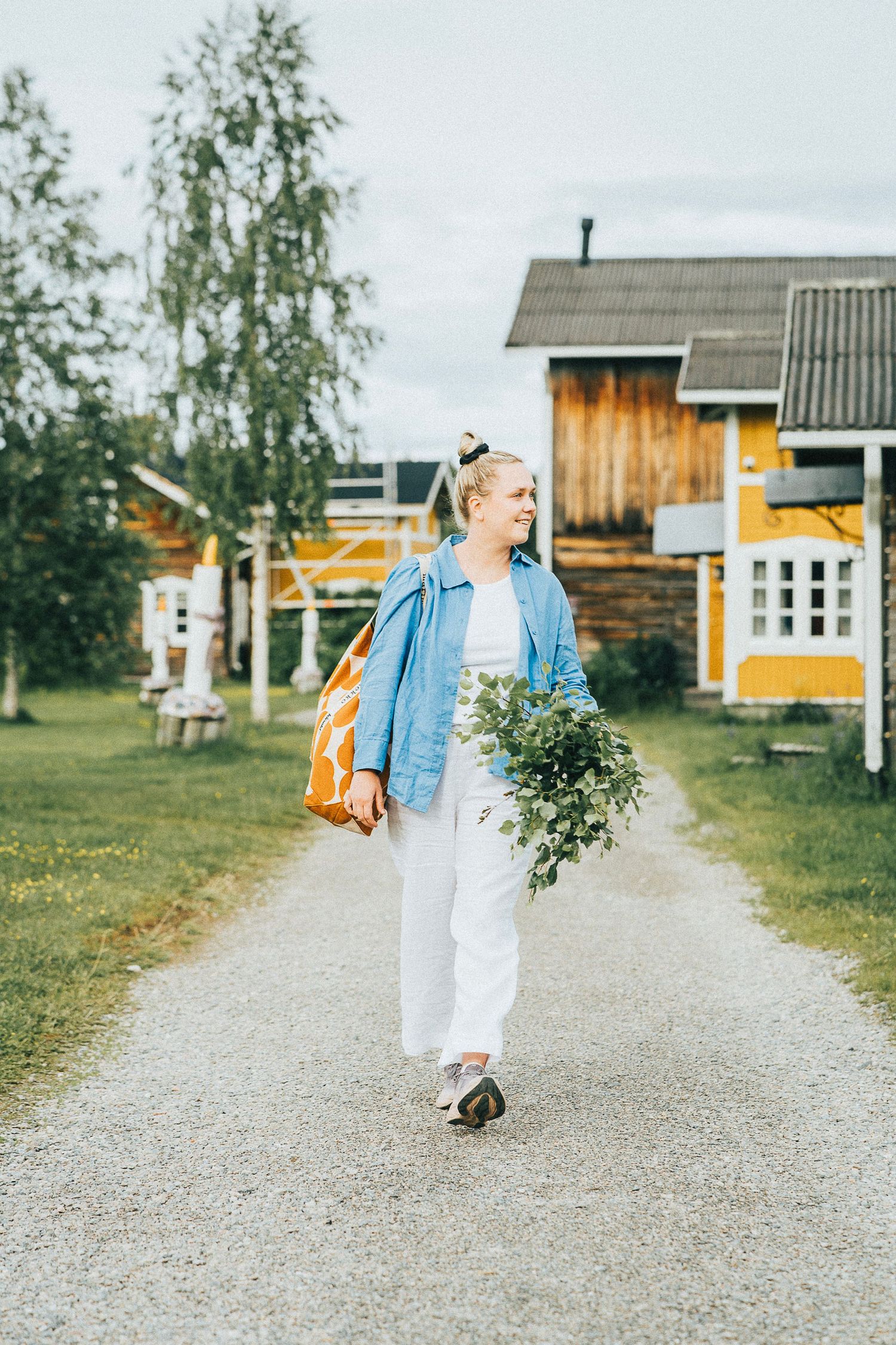 Woman walking on gravel path holding greenery bouquet and orange bag near yellow wooden houses.