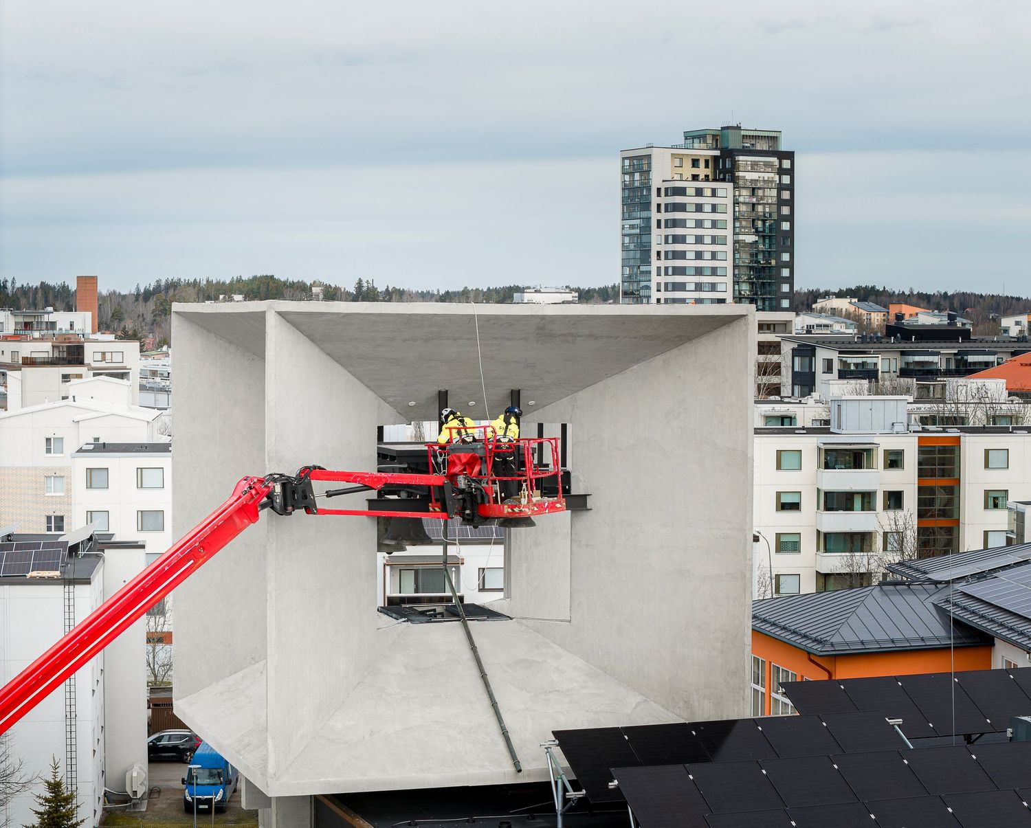 Workers on a red aerial lift platform inspect a large concrete structure on a rooftop in an urban cityscape.