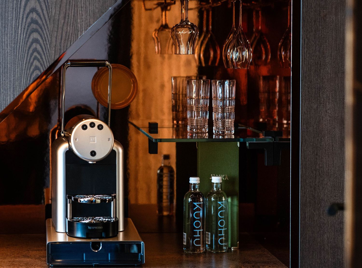 Nespresso coffee machine beside an open wooden cabinet with glassware and green beverage bottles.