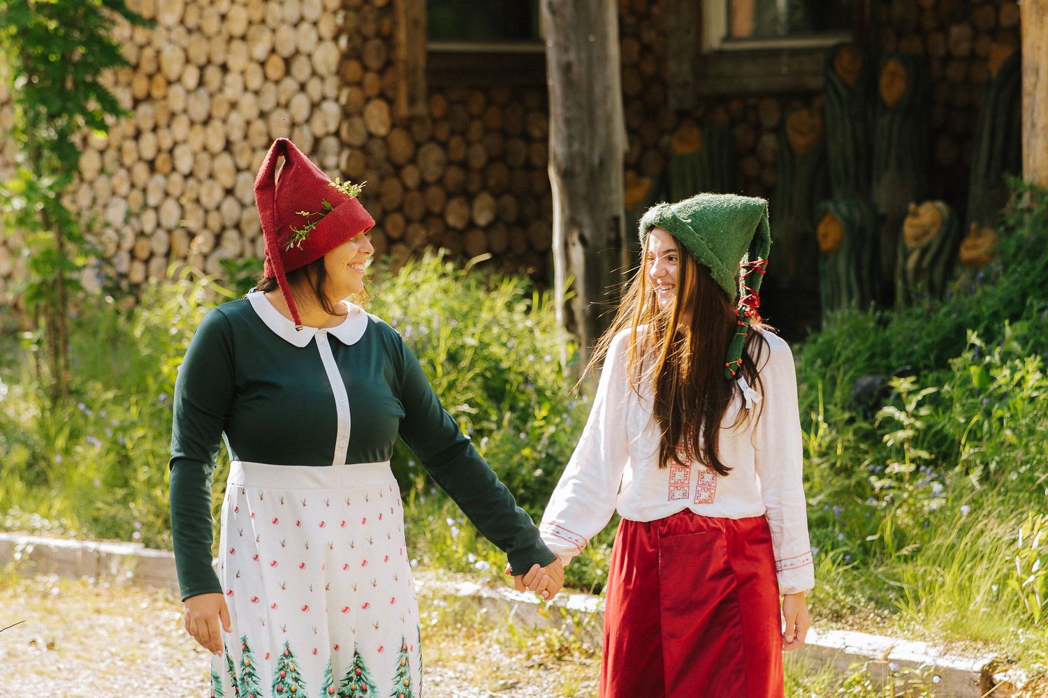 Two women in traditional folk costumes with red elf hats hold hands outdoors near a rustic wooden building.