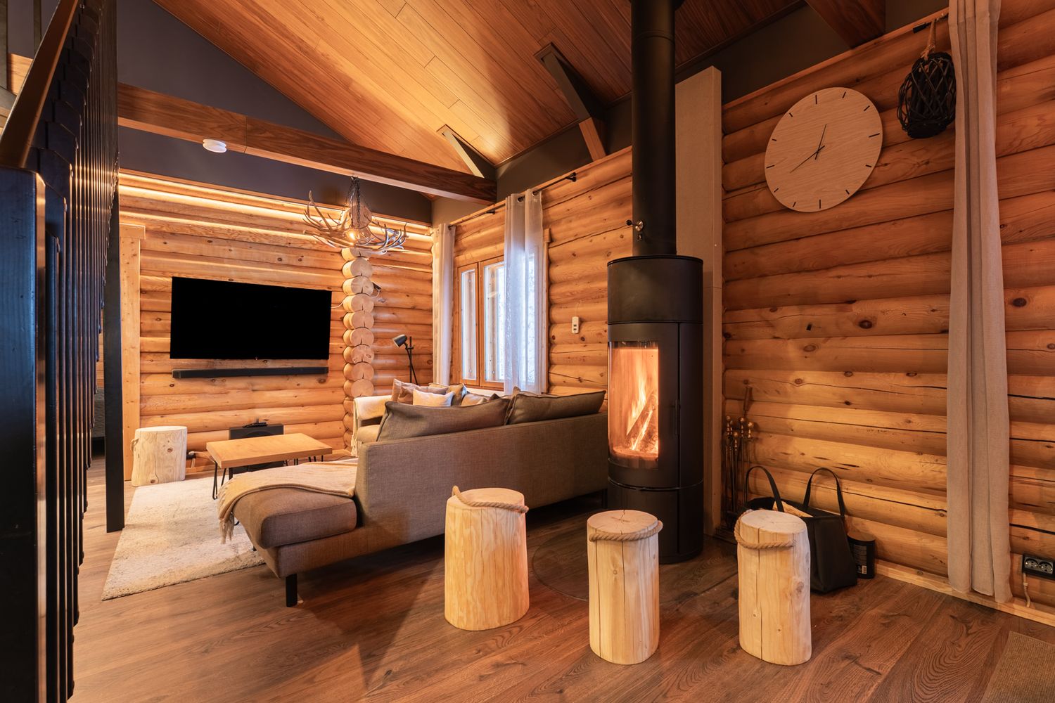Cozy log cabin living room with wood-burning stove, rustic wooden stools, flat-screen TV, and warm ambient lighting.