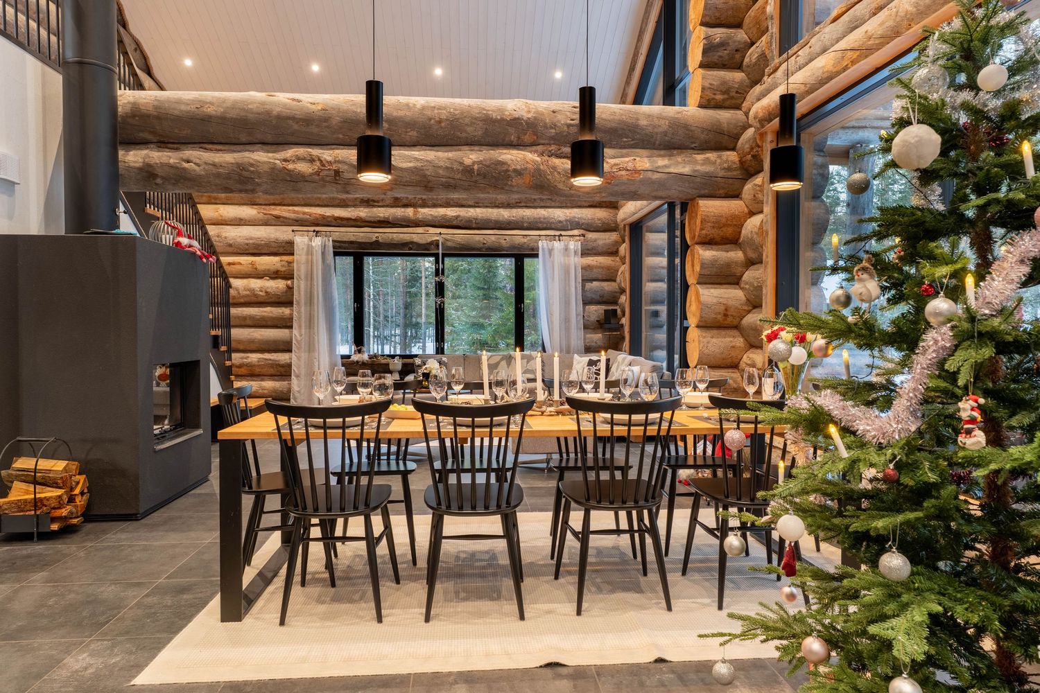 Log cabin dining area with long table, black chairs, Christmas tree, fireplace, and winter forest backdrop.