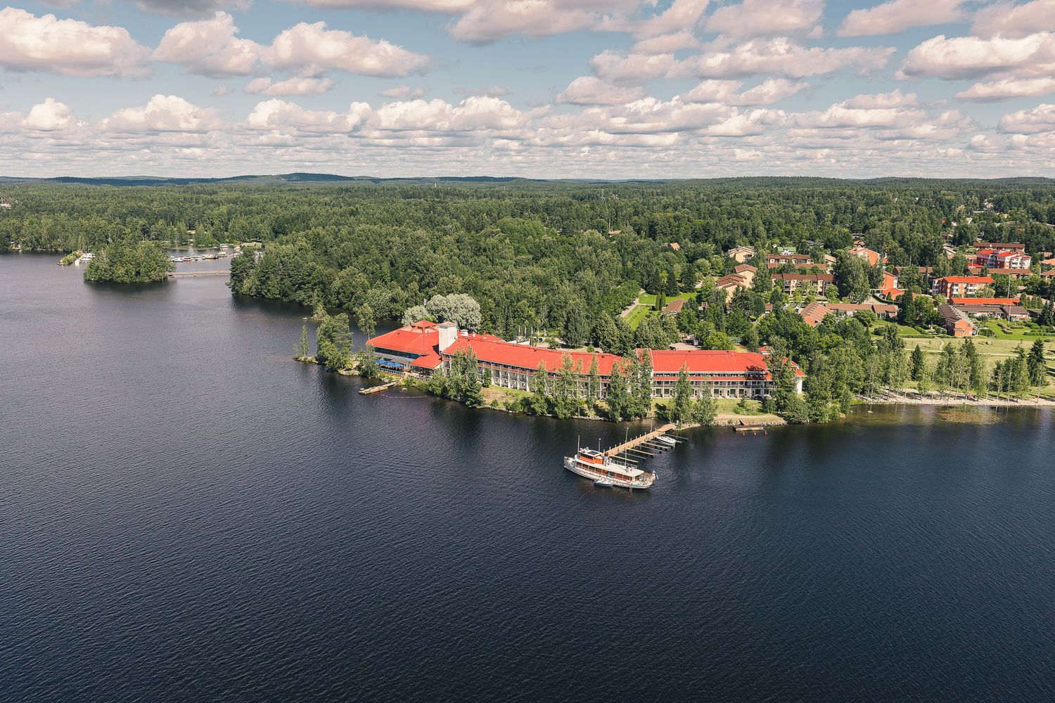 Aerial view of a lakeside resort with red-roofed buildings surrounded by lush green forest in Sweden.