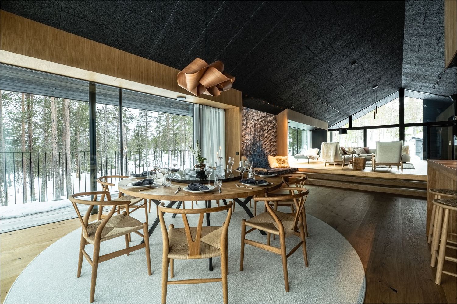 Modern Scandinavian dining room with large windows, oval black table, wooden wishbone chairs, and copper pendant light.