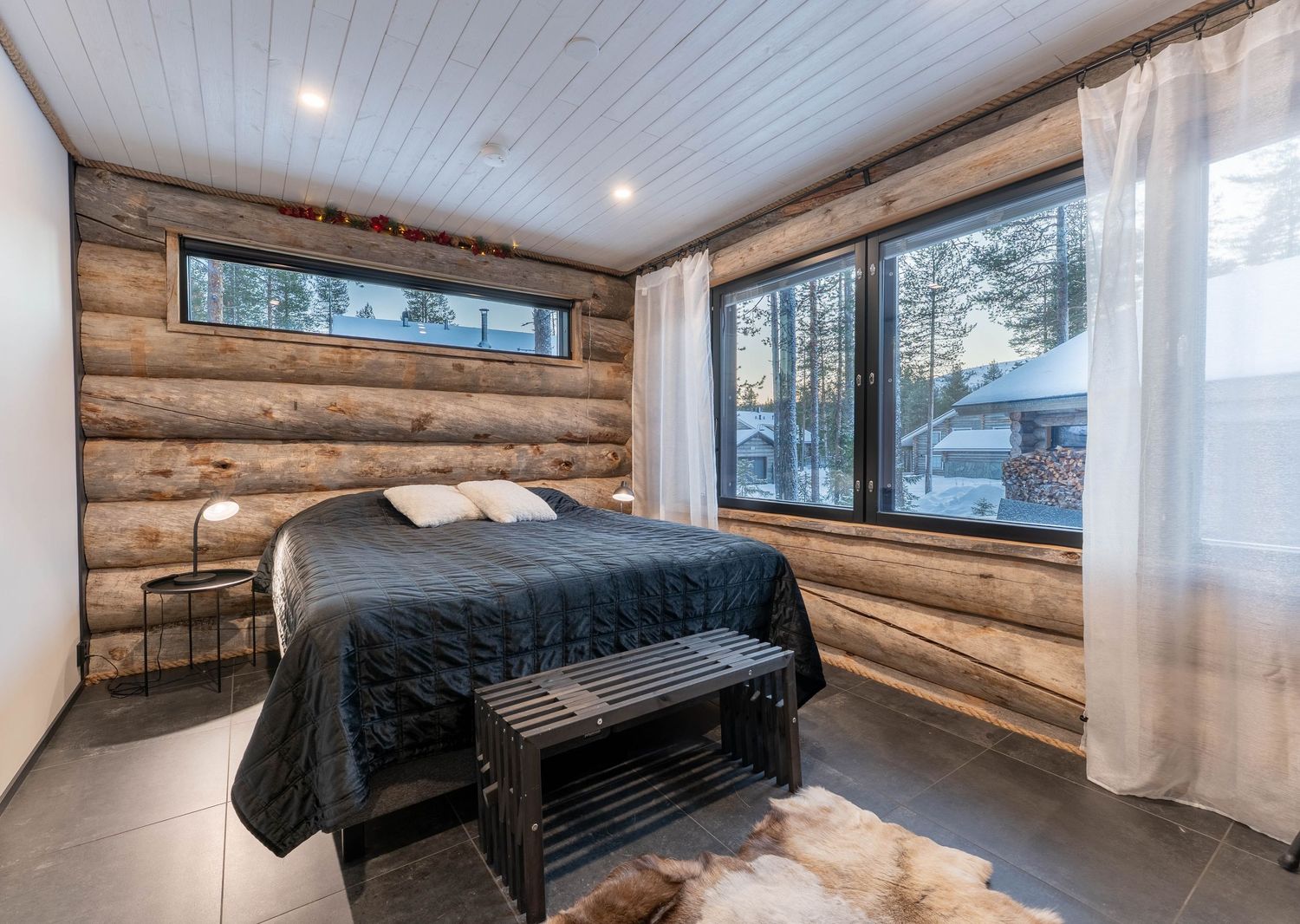 Cozy log cabin bedroom with rustic wood walls, dark bedding, bench, and sheer curtains over winter forest windows.