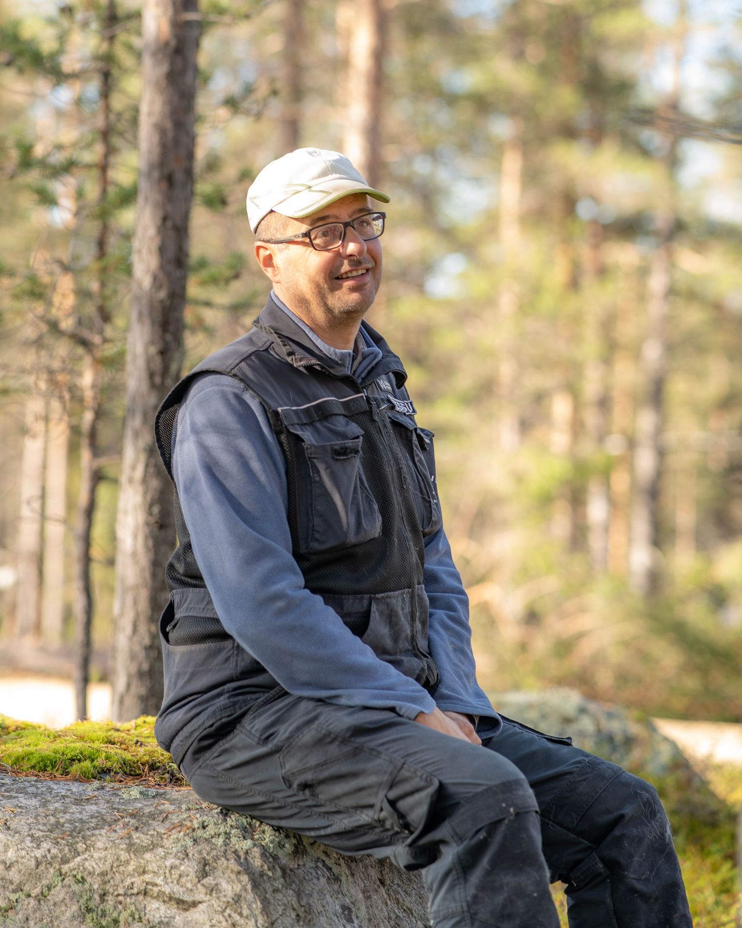 Man in glasses and cap sitting on a mossy rock in a sunlit forest, wearing a black vest over blue clothing.