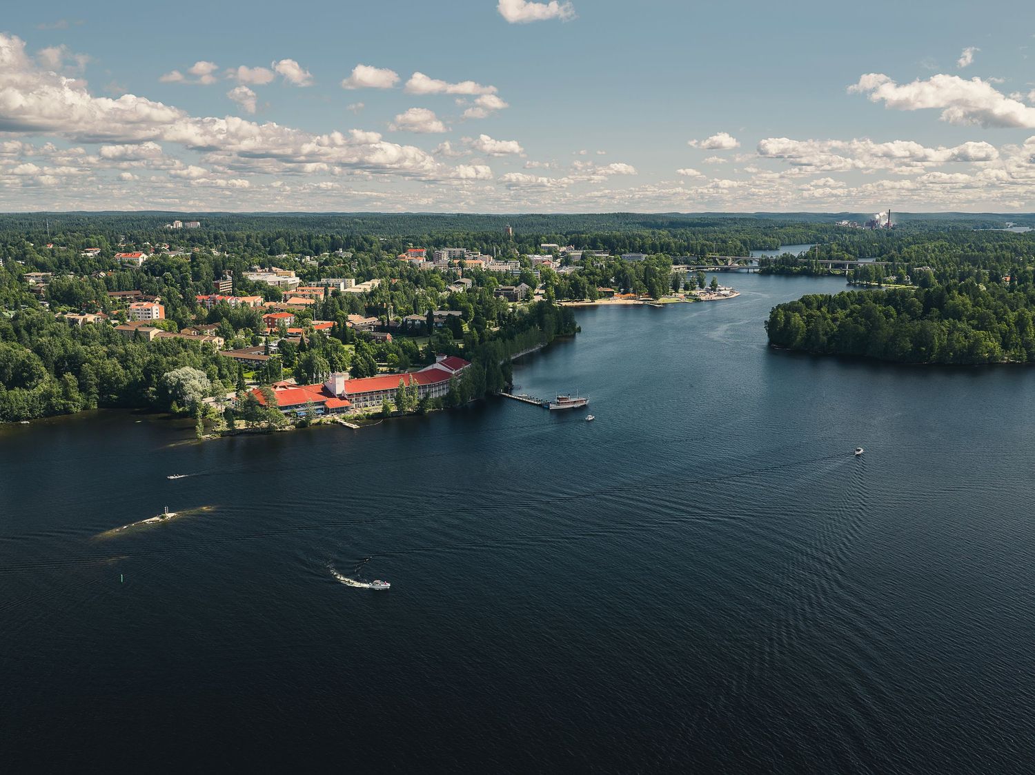Aerial view of a lakeside town surrounded by lush green forests and calm blue waters under a partly cloudy sky.
