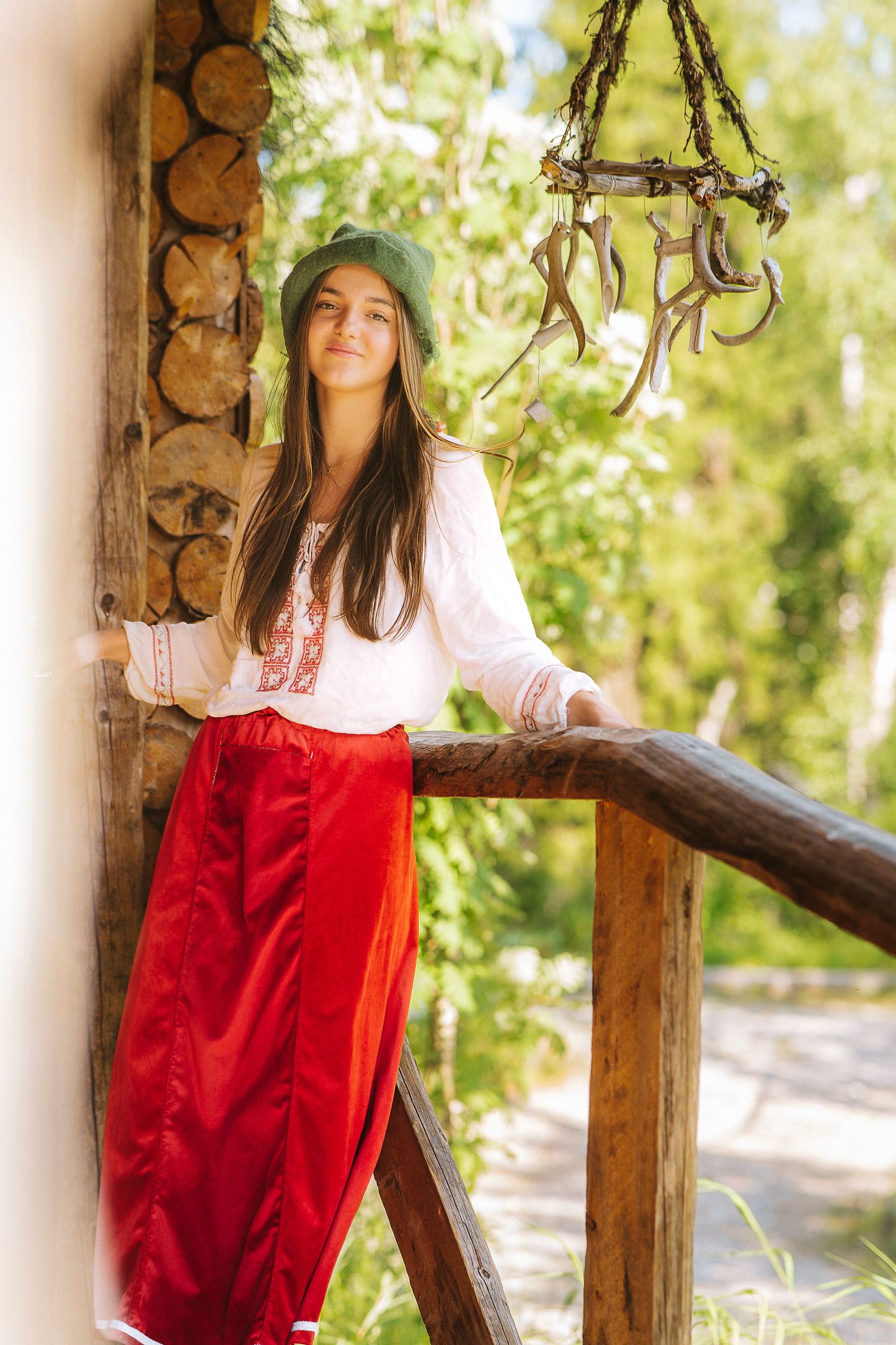 Young woman in traditional red skirt and white embroidered blouse with green hat poses on rustic wooden porch.