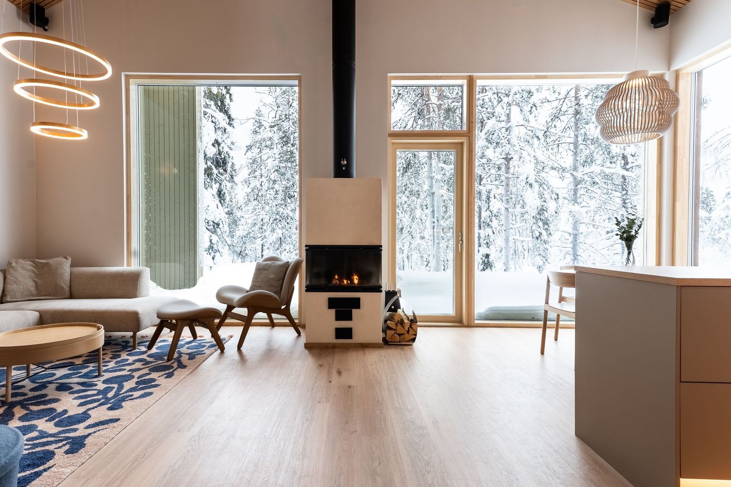 Bright Scandinavian living space with corner fireplace, panoramic snow-covered forest windows, and blue rug.