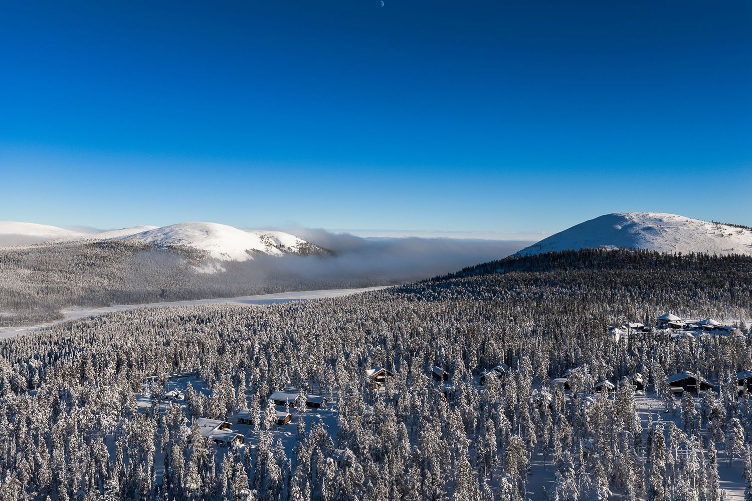 Aerial winter panorama of snow-covered pine forest with mountain peaks under a clear blue sky in Lapland.