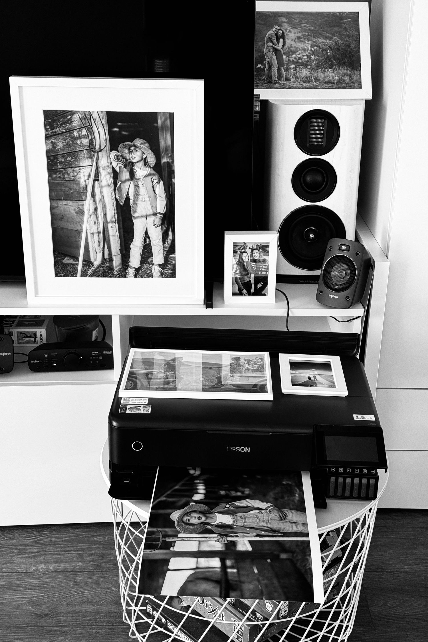 A black and white photo display setup with framed prints and a printer on a wire basket stand.