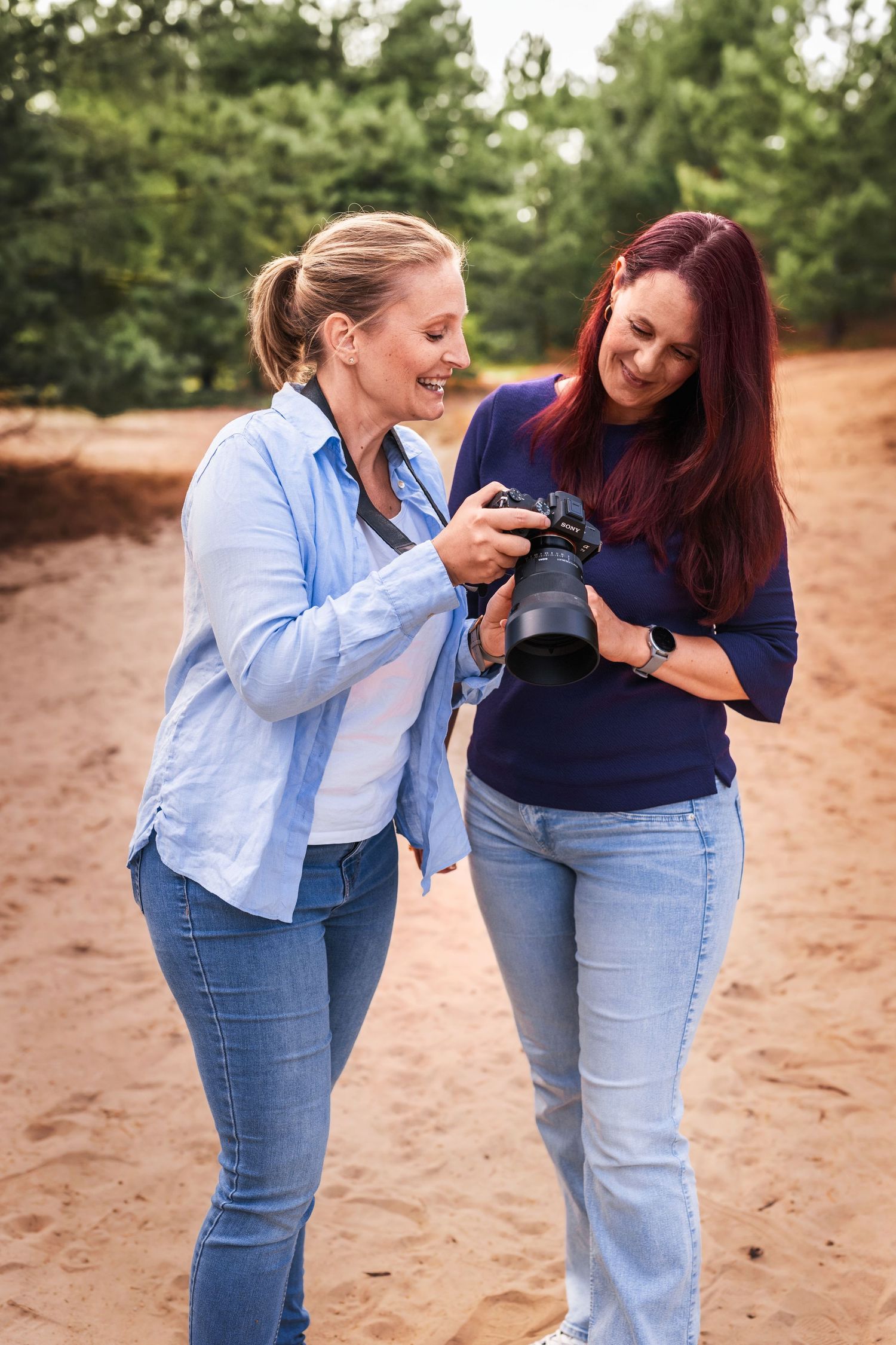 Two women smile while reviewing photos on a camera outdoors on a sandy forest path.